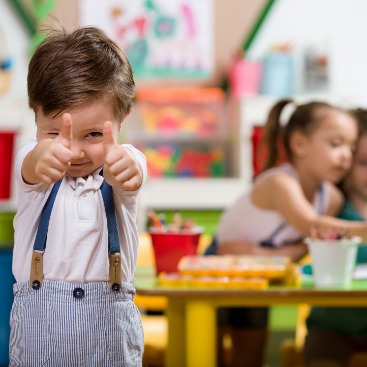 Kachel: Kindergarten Junge gibt Thumbs Up, im Hintergrund Kinder am Tisch