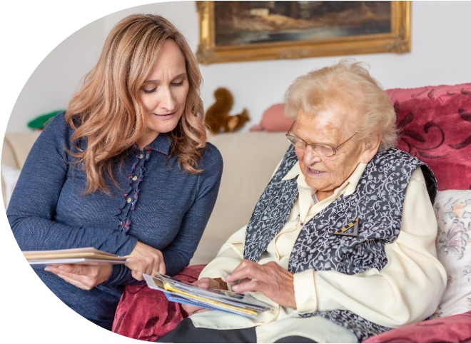 Eine jüngere Frau sitzt mit einer Seniorin auf einem Sofa. Sie sehen sich Fotos an.