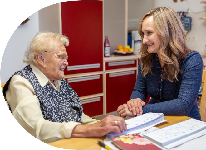 Eine Seniorin und eine junge Frau sitzen gemeinsam an einem Tisch und lächeln sich an. Vor sich haben sie einen Terminkalender und verschiedene Papiere ausgebreitet