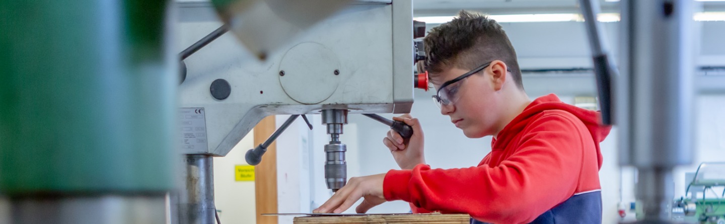 Header: Jugendlicher Schüler bohrt in einer Werkstatt an einer Standbohrmaschine ein Metallstück