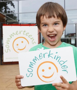 Sommerkinder: Ein Junge hält das Sommerkinderlogo auf einem DinA 4 Blatt und Lacht dabei