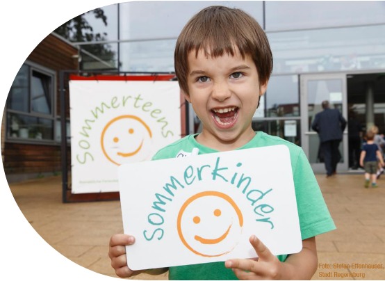 Sommerkinder: Ein Junge hält das Sommerkinderlogo auf einem DinA 4 Blatt und Lacht dabei