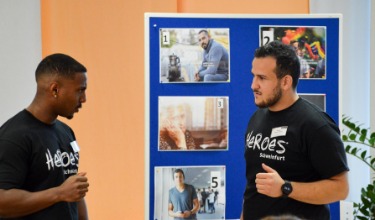HEROES Yuel Emha und Hasan Sayed Yahya leiten einen Workshop am HEROES Fachtag in Schweinfurt. Sie stehen vor einer Pinnwand mit Fotos.