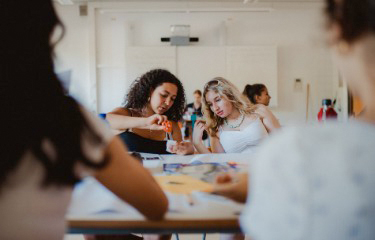 Jugendliche sitzen im Klassenraum am Tisch, 2 Mädchen im Hintergrund